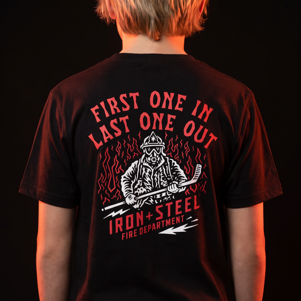 First One In Last One Out Hockey Tee in black worn by a young hockey player, back print of a firefighter holding a hockey stick in front of flames with an Iron+Steel Fire Department banner