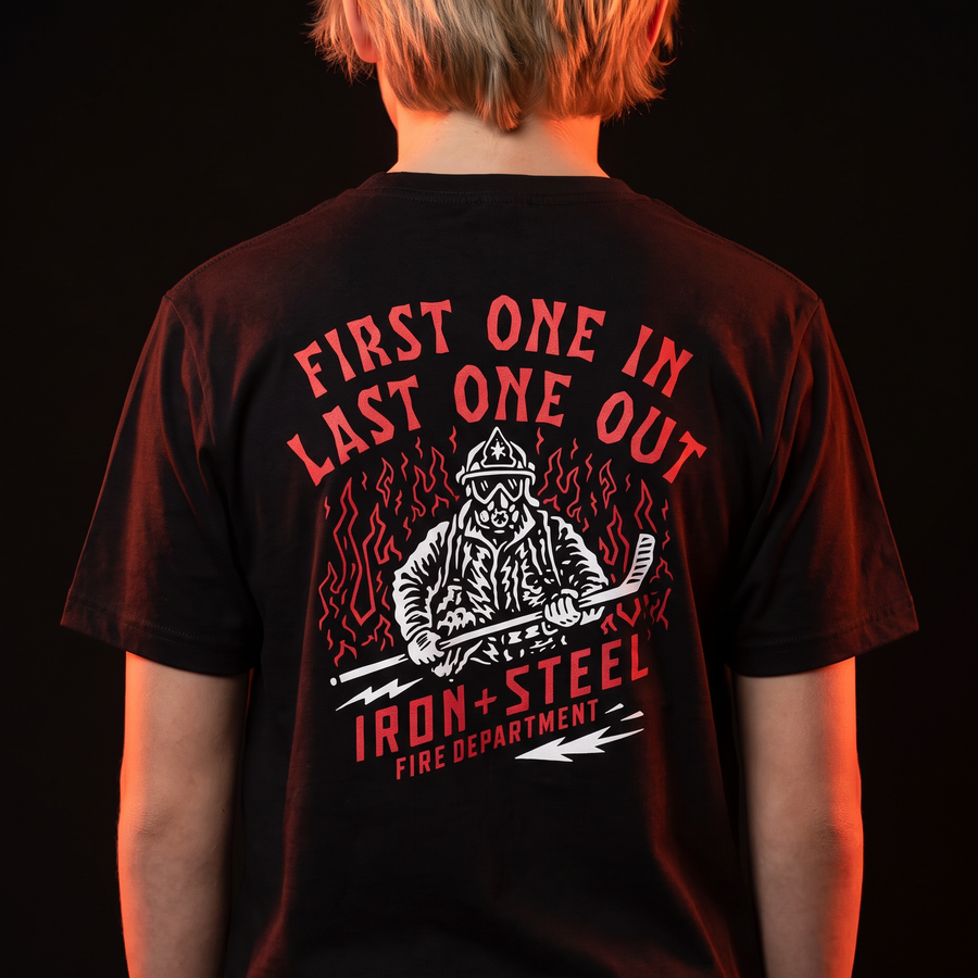 First One In Last One Out Hockey Tee in black worn by a young hockey player, back print of a firefighter holding a hockey stick in front of flames with an Iron+Steel Fire Department banner