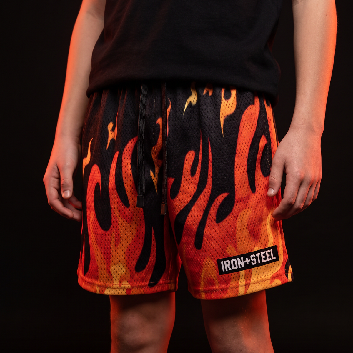 Flames Hockey Mesh Shorts worn by a young hockey player, black mesh with orange and red flame print and IRON+STEEL hem tag
