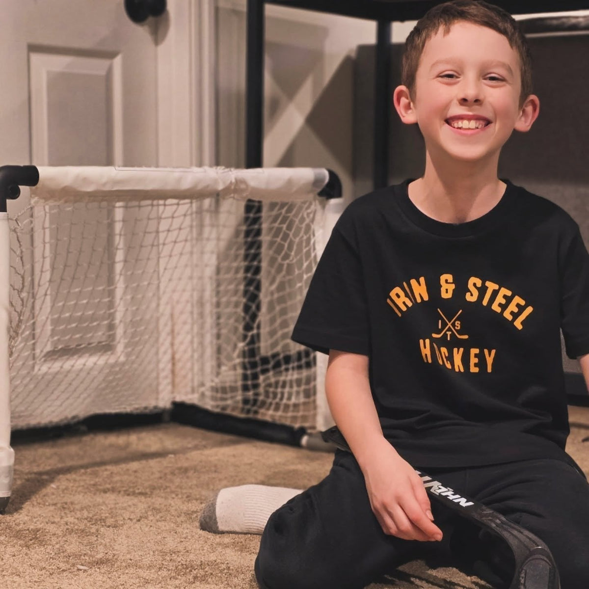 Smiling boy with a mini stick beside a basement hockey net in an Iron+Steel tee