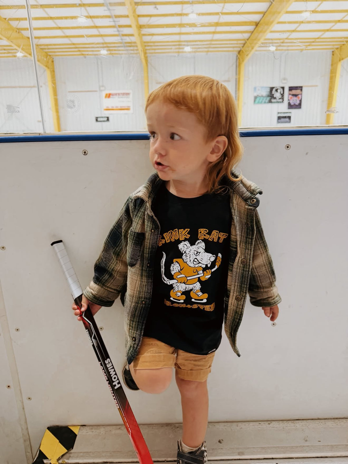 Toddler with a hockey stick at the boards in an Iron+Steel Rink Rat tee