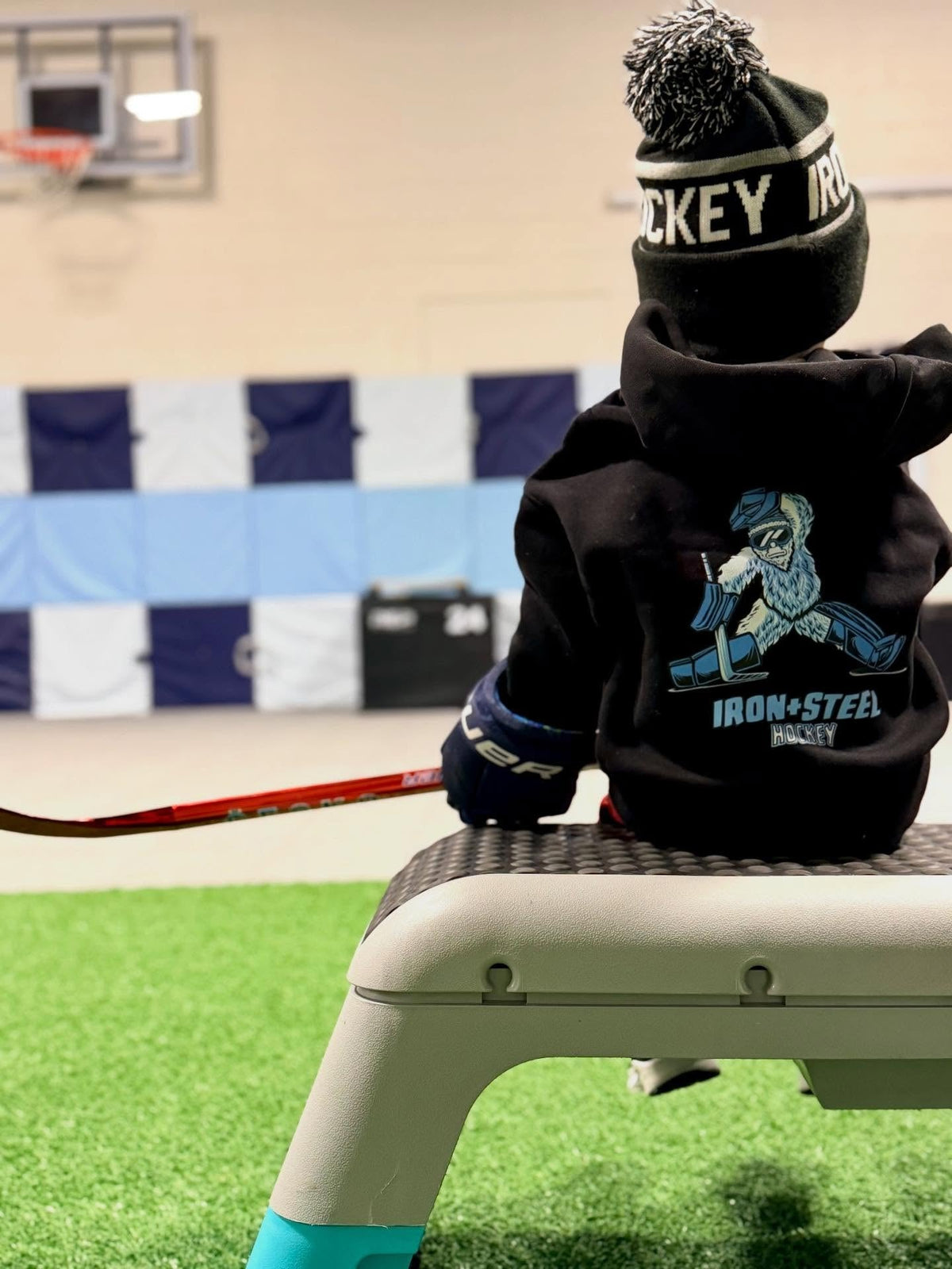 Young player on a bench in an Iron+Steel goalie hoodie and hockey beanie